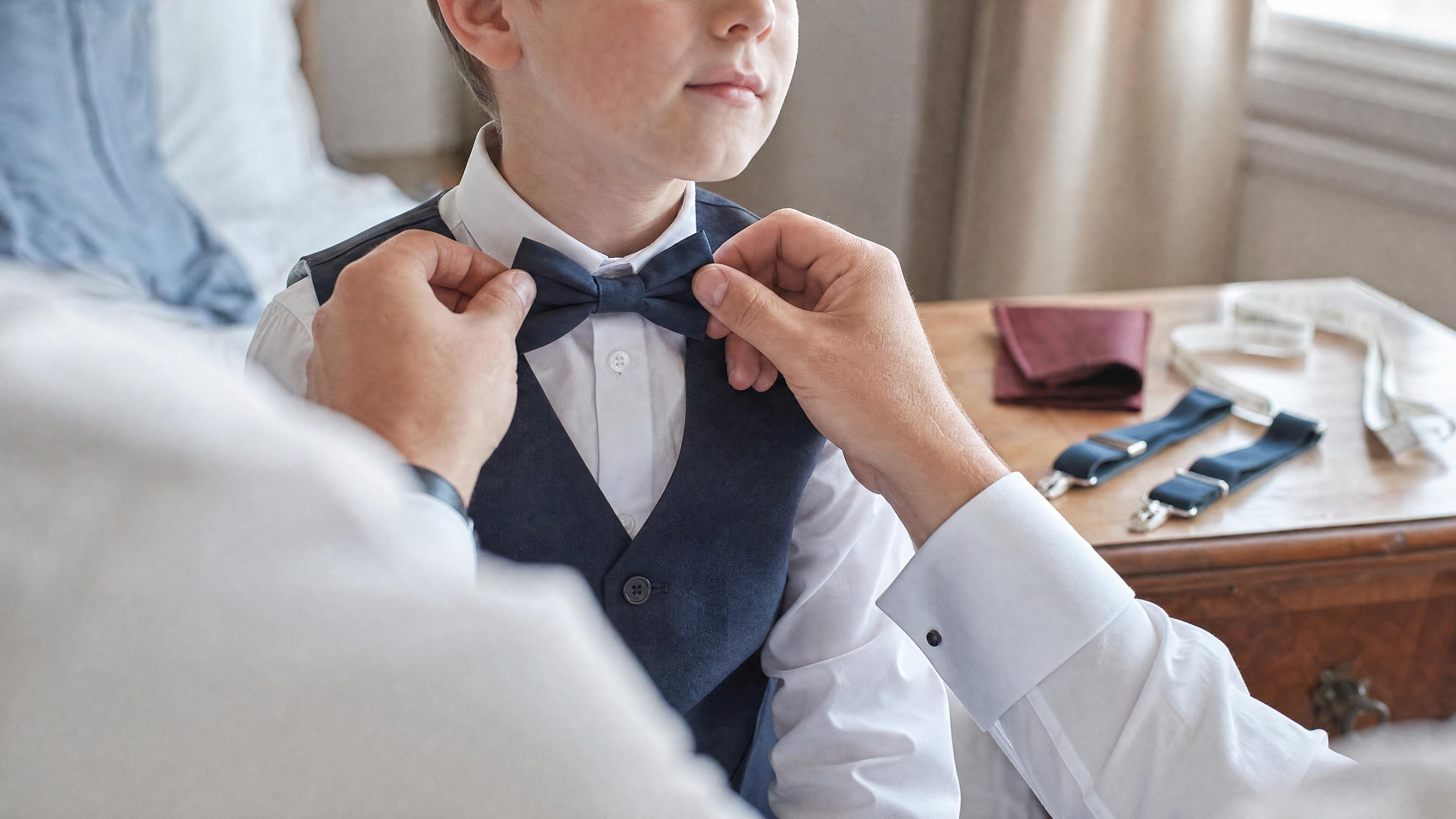 Un adulte ajuste un nœud papillon sur un enfant en chemise blanche, avec un mètre ruban visible sur une commode, dans une chambre lumineuse avant une cérémonie Obraz